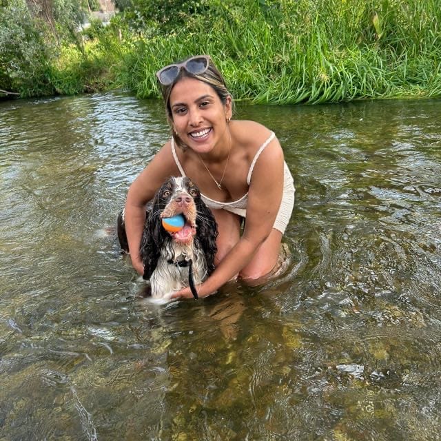 Shivani and a dog playing with a ball in the water in a wooded area